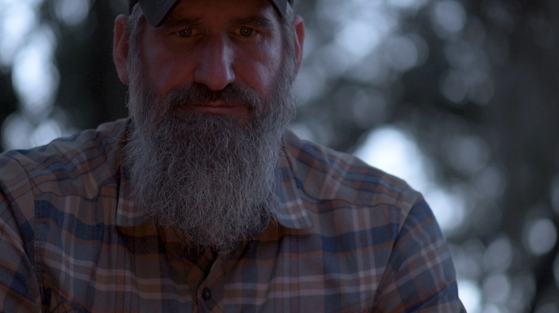 Survival instructor Joshua Enyart staring at the ground in deep thought beside a campfire at night, firelight reflecting off his skin — illustrating the psychological and mental dimension of wilderness survival