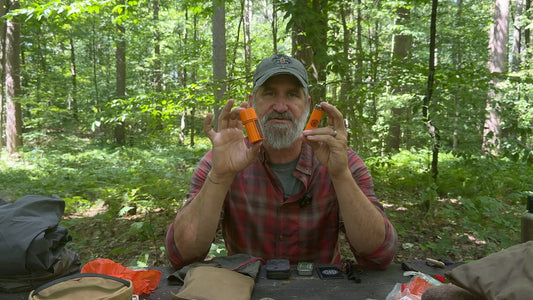 Joshua Enyart reviewing wilderness survival gear components at a field table in the woods — survival kit gear review
