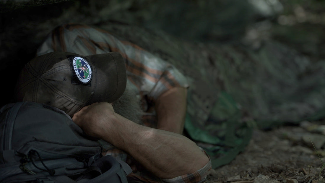 Joshua Enyart lies prone in a low-profile concealed bug out shelter, demonstrating BLISS signature reduction on the forest floor.