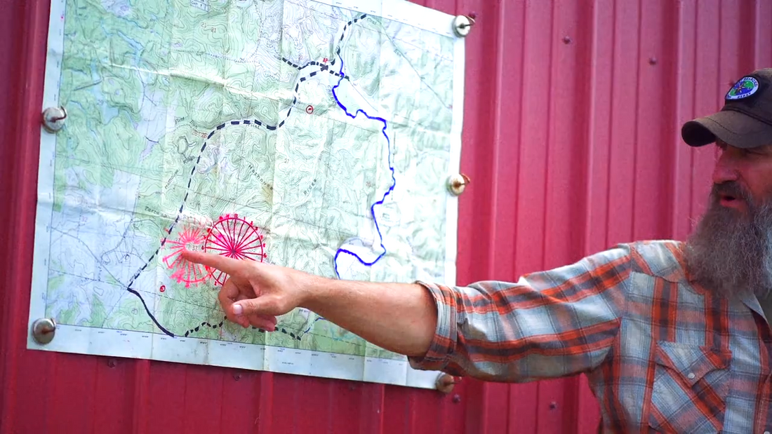 Joshua Enyart stands in front of a wall-mounted topographic map, pointing at a plotted navigation corridor — cross-country bug out routing demonstration.