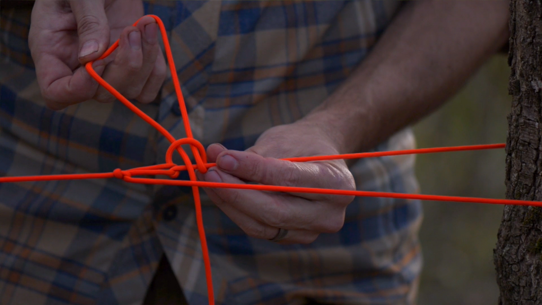 Tying a modified trucker's hitch knot with orange paracord tied to a tree — essential wilderness survival knot technique