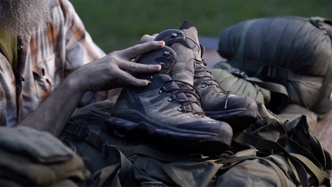 Close-up of hands inspecting a pair of worn Salomon hiking boots resting on top of a loaded bug out pack at dusk.