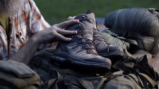 Close-up of hands inspecting a pair of worn Salomon hiking boots resting on top of a loaded bug out pack at dusk.