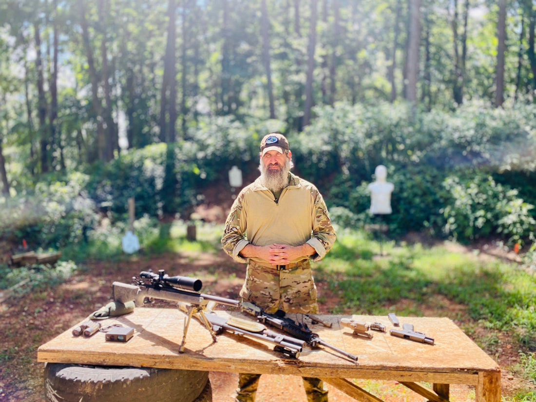 Joshua Enyart stands behind a wooden range table holding a scoped rifle; additional firearms, magazines, and gear staged in front of him.