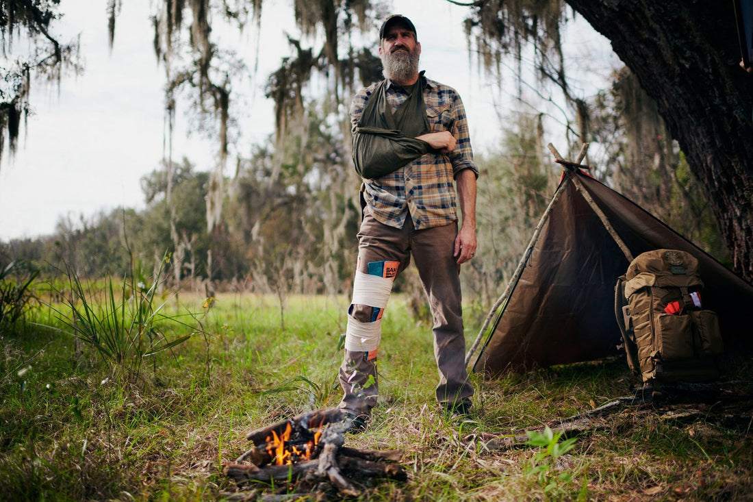 Special Forces veteran Joshua Enyart in the field with his arm in a sling and knee immobilized, seated beside a campfire in front of a survival shelter — illustrating the stay-or-go decision when injury changes the calculus