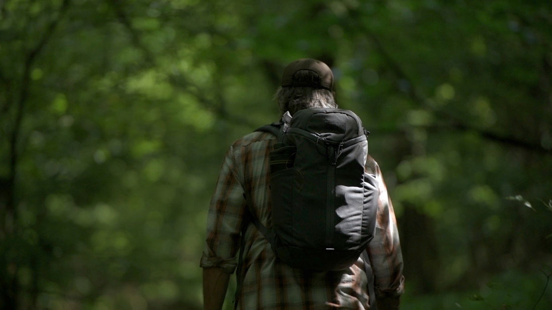Bearded man in plaid shirt and ball cap, carrying a loaded black backpack, walks away from the camera into dense green woods.