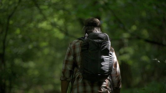 Bearded man in plaid shirt and ball cap, carrying a loaded black backpack, walks away from the camera into dense green woods.