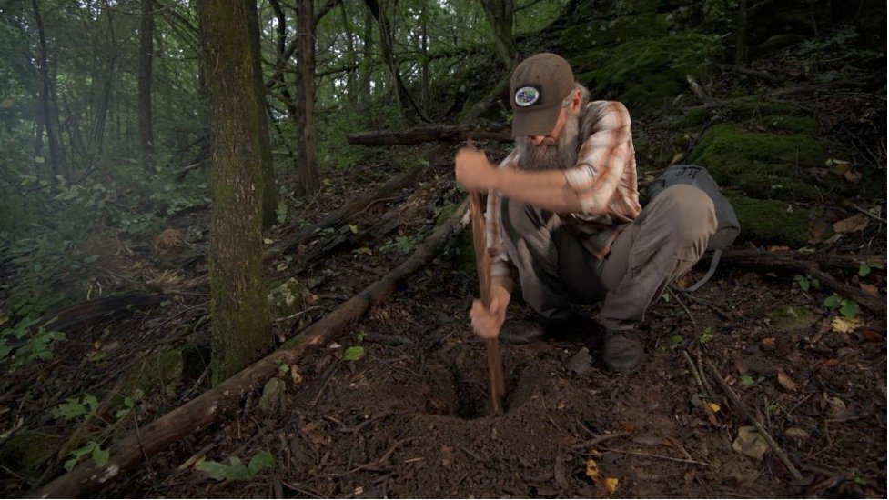 Joshua Enyart kneels in a wooded ravine, using a stick to dig the air-intake hole of a Dakota fire pit.