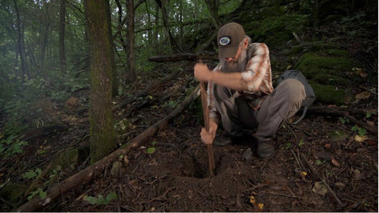 Joshua Enyart kneels in a wooded ravine, using a stick to dig the air-intake hole of a Dakota fire pit.