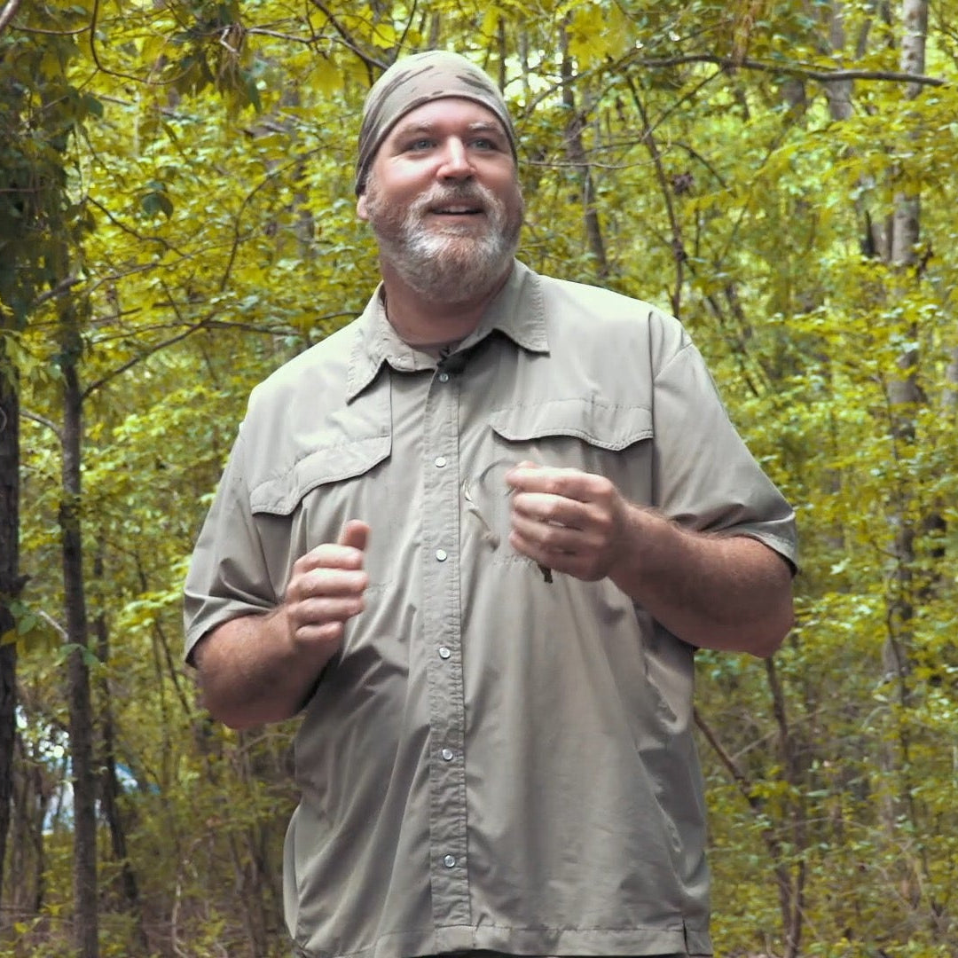Man standing in a forest wearing a beige shirt and cap.