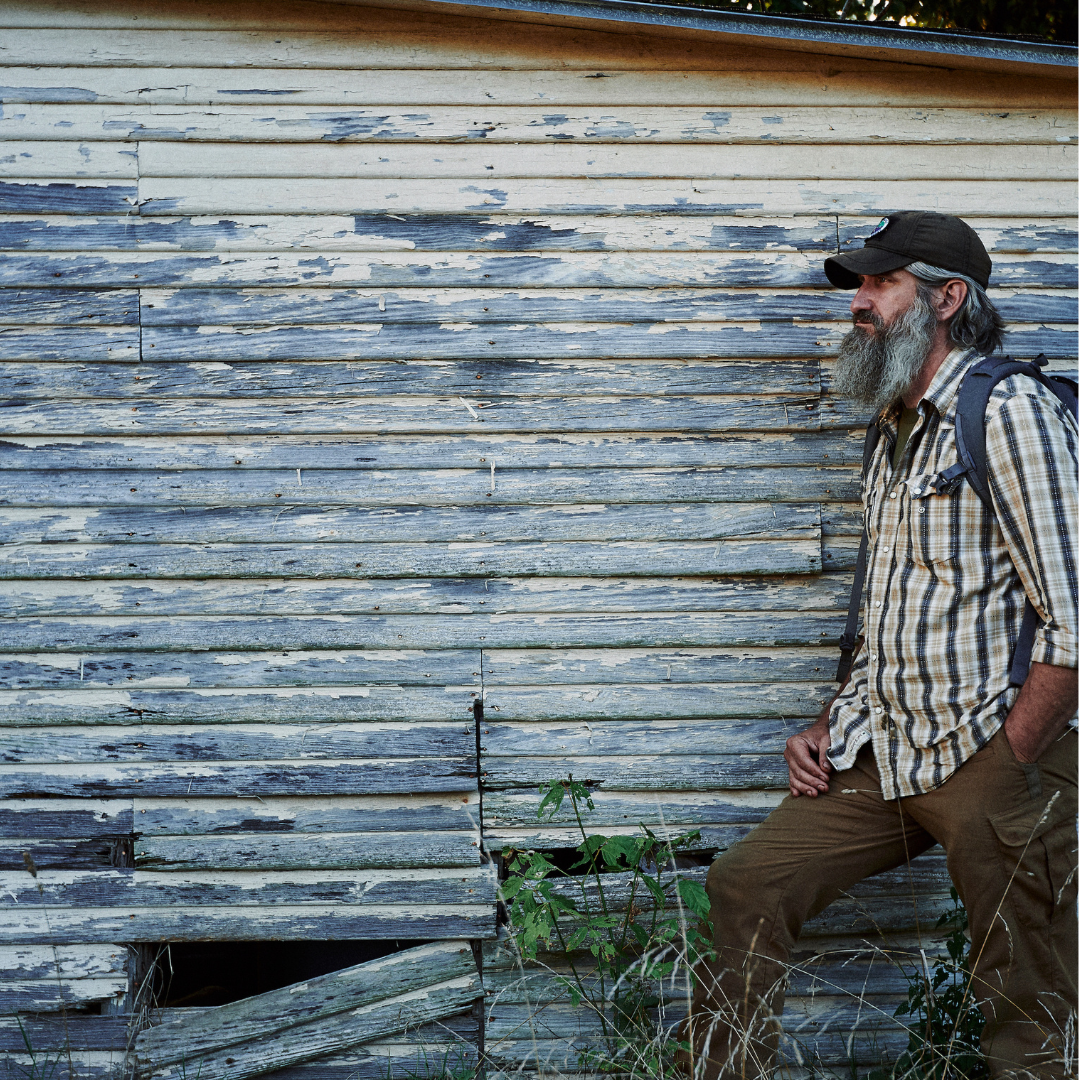 Man with a beard and hat leaning against a rustic wooden wall